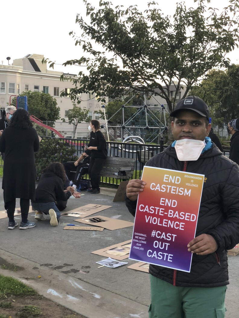 A man holds a sign at a protest. Behind him, four other people prepare signs. A playground looms in the background.