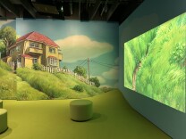 A corner of a room with grass-green carpet and round stools in the same color. The left wall is covered in a mural of a yellow home on a grassy hill. The right wall is showing a clip from the movie Ponyo featuring bright green animated grass.