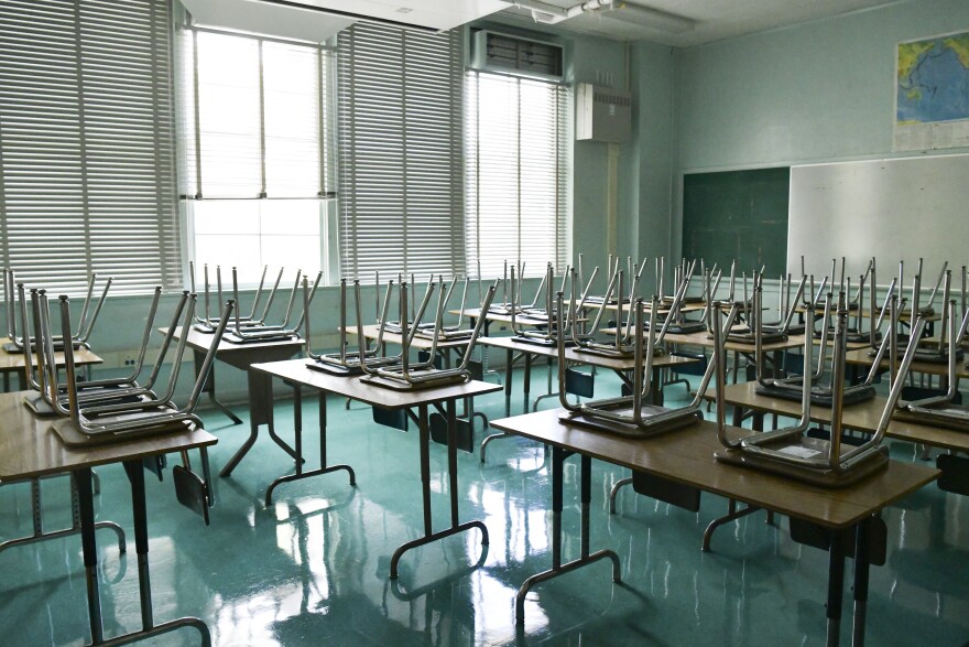 Two chairs per desk are resting seat down on the desktop in an empty classoom. The floors are green and reflect the light.