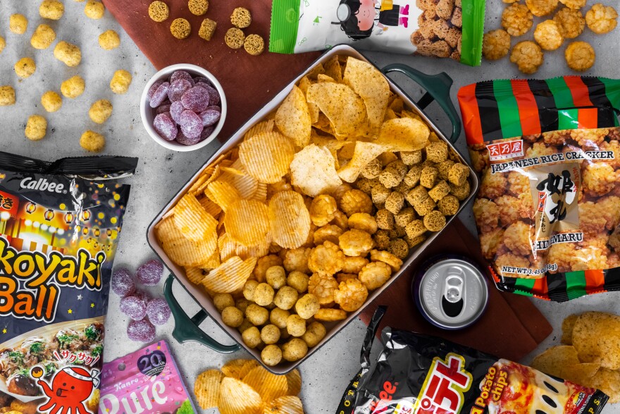 A variety of brightly colored snack bags and snacks are laid out on a table, surrounding a large ceramic container containing different-shaped chips. There is also an open beverage can next to the container and a small bowl of purple gummy candy