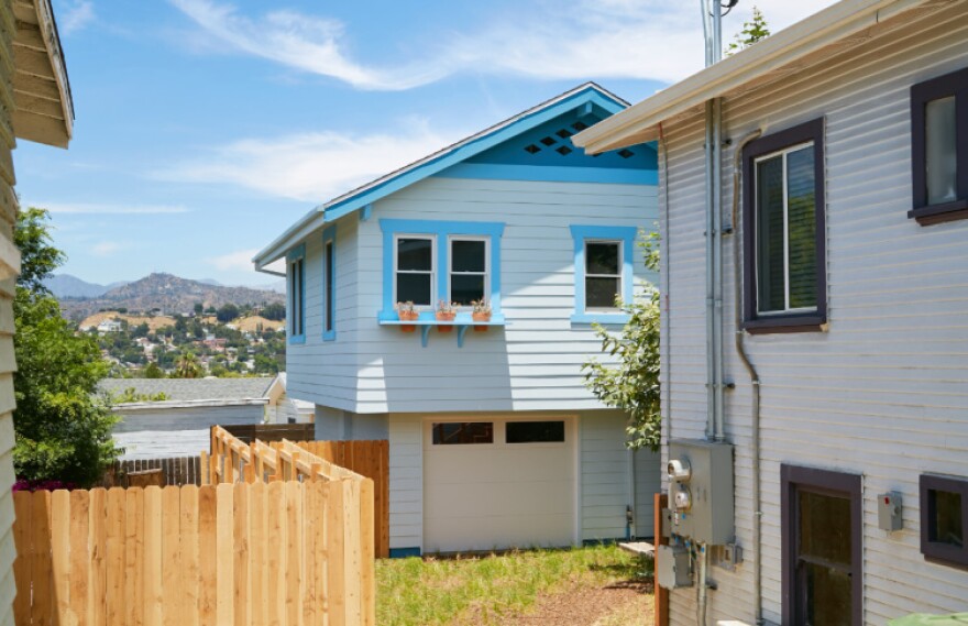 A two-floor house with a garage on the back topped by a small house painted in white and light blue, surrounded by a wooden fence.