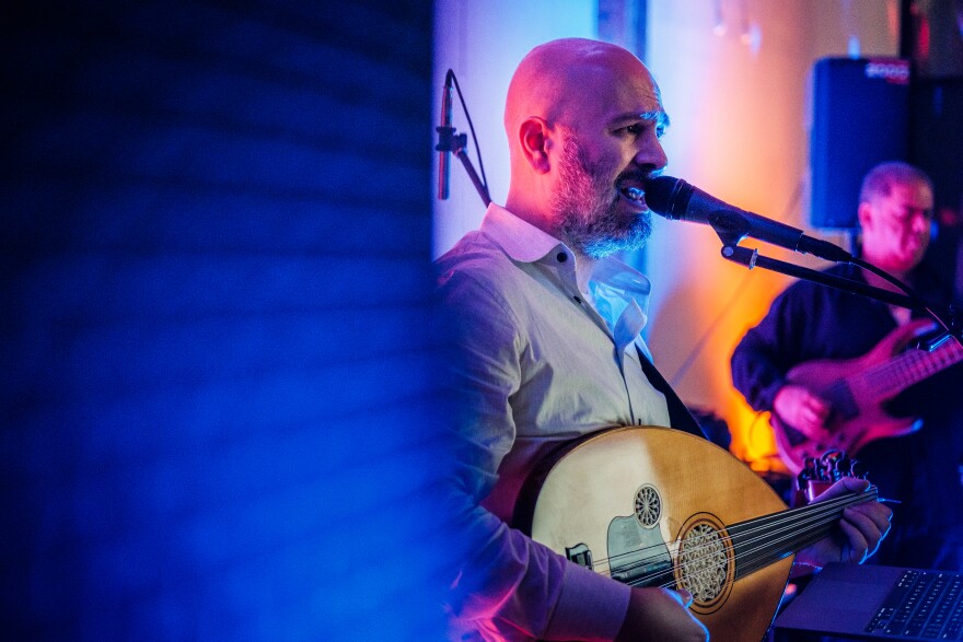A fair skinned man who is bald with a beard holds an a stringed instrument known as an oud while he sings into a microphone. There is another man behind him slightly out of focus, playing a bass guitar wearing a dark colored shirt.