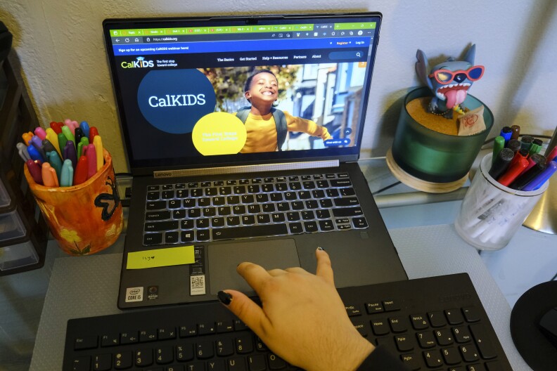 A black laptop displays the CalKIDS homepage. To the left there is an orange ceramic cup of colorful permanent markers and to the right, a plastic figure of the cartoon character Stitch.