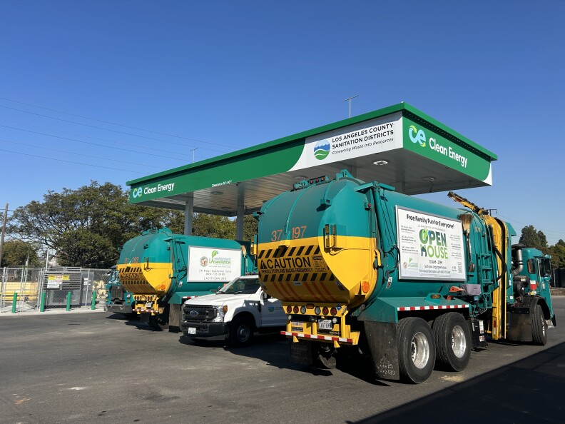 A wide shot of green trash trucks refueling at a CNG station. The shade platform over the fueling station is green and reads "clean energy"  and "Los Angeles County Sanitation Districts." It's sunny under a clear blue sky. 