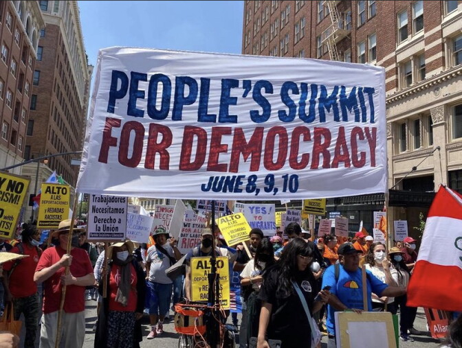 Among a crowd of people, a large white flowing banner in the air held by two people reads, "The People's Summit For Democracy," in blue and red, while other poster and picket signs are waving in the air around it