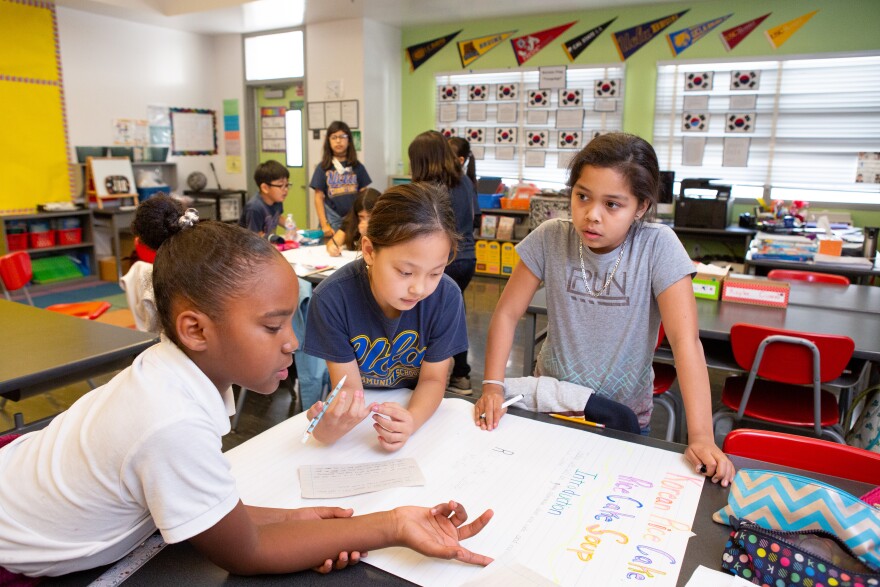 Three female elementary students lean on a table in a classroom while working on a poster. The classroom wall behind them is painted green, and above the windows runs a line of pennants. Other students are working in the background working on a project. The words "Rice Cake Soup" and "Introduction" can be seen on the poster.