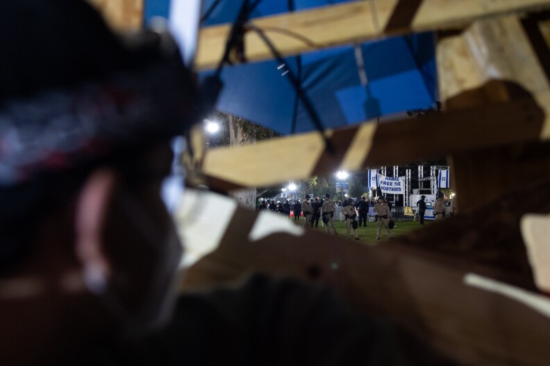 A person peaks through an opening in a makeshift wooden barrier at officers who stand in a lawn.