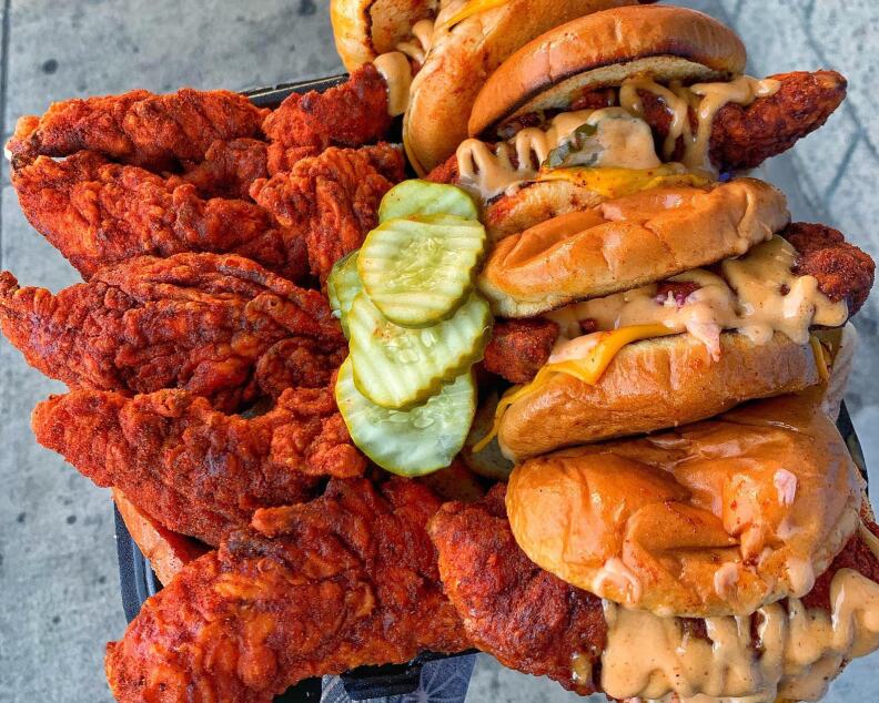 A platter showing orange fried chicken tenders next to a mound of fried chicken buns, all doused in a light tan sauce; in the middle is a line of pickles.