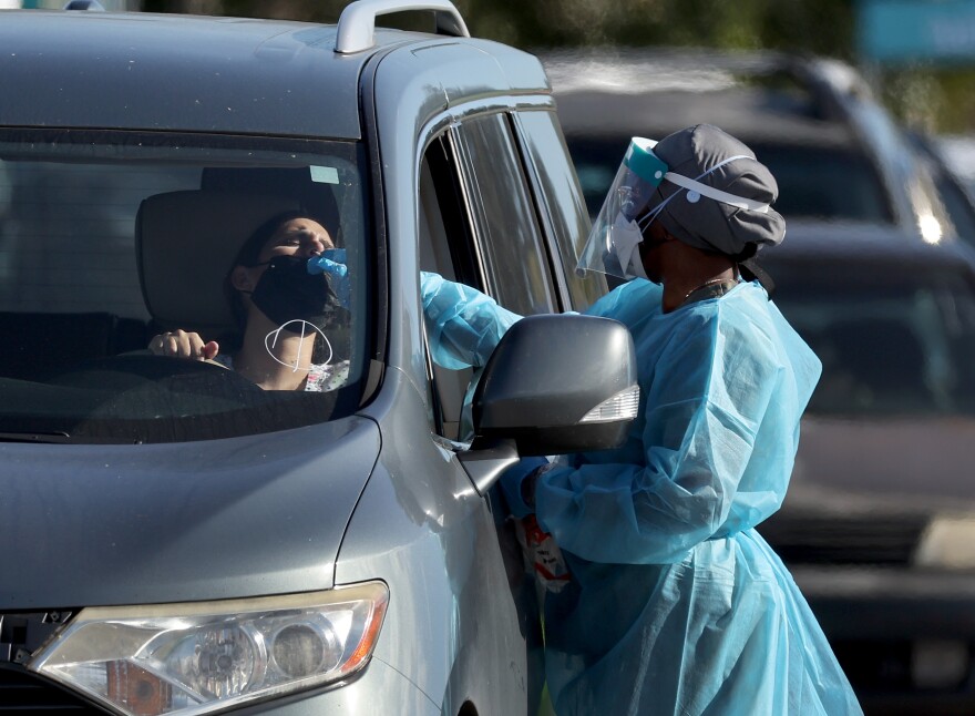 A health care worker wearing blue protective equipment over their scrubs and a mask and face shield conducts a test with a person in a gray vehicle. There is a line of cars in the background.