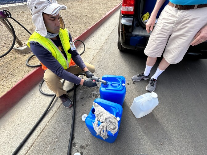 A person with a yellow vest on crouches down to fill up small blue containers with a hose that shoots out water.