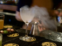 Smoke swirls above a stemmed cocktail glass on a bar mat, next to a metal jigger and a dish of limes.