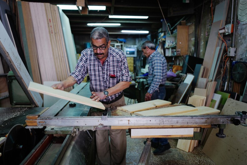 Two older men in plaid shirts work with wood boards.
