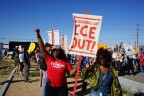 A Black woman wearing a baseball cap and sunglasses wears a red shirt that says "My Tribe Rise." Her right fist is raised and in her left hand she holds a large white sign that says "Neighbors Say ICE OUT!" She stands next to a dark skin-tined woman with medium-length dark hair who wears sunglasses and is making her right hand into a peace sign.
