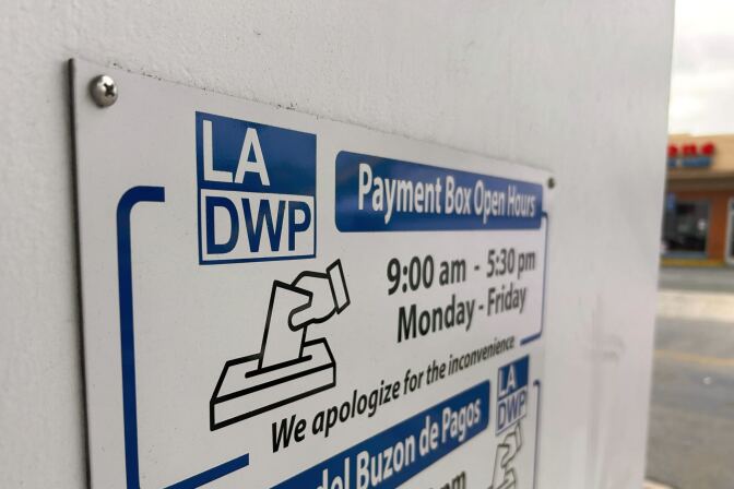 A close up of a payment drop box sign on the white wall of the LADWP building. It says in blue, black and white the LADWP payment box open hours with an icon of a hand putting an evelope into a box cutout. The box is open 9am to 5:30pm Monday through Friday. Below it has the words in Spanish.