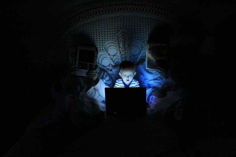 Small child sitting in bed with parents looking a screen. Everything is dark except for the child's face only brighten from the screen.