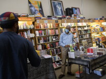 A Black man with grey hair stands in front of a display table in a bookstore, holding a copy of a book featuring President Barack Obama on the cover. The man is dressed in a light blue shirt, khakis and a blue surgical face mask. Walls of books are behind him. 