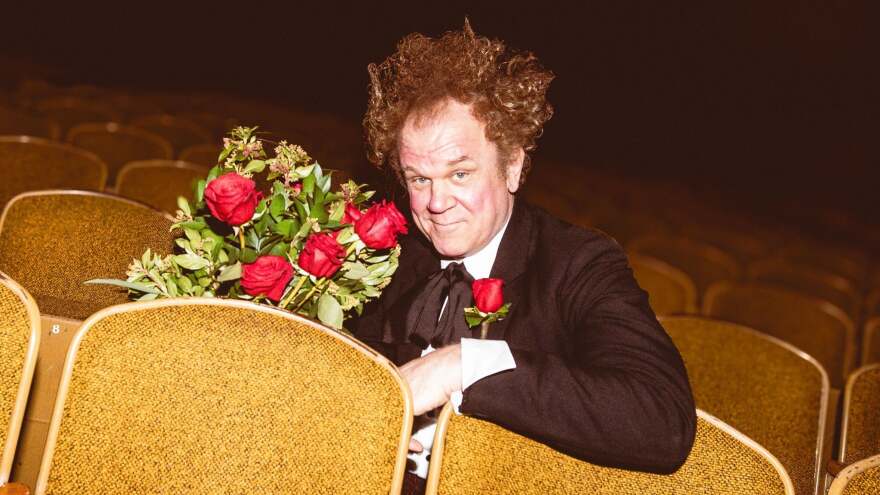 John C. Reilly, wearing a suit, holds roses and smirks at the camera as he sits in yellow theater seats. He's in character as Mister Romantic.