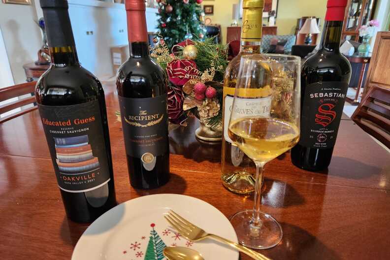 A festive scene with a Christmas tree in the background; in the foreground there is a white plate with a painted Christmas tree and a gold fork placed on top. Next to it is a wine glass with yellowy white wine in it, surrounded by four bottles of wine, three red, one white