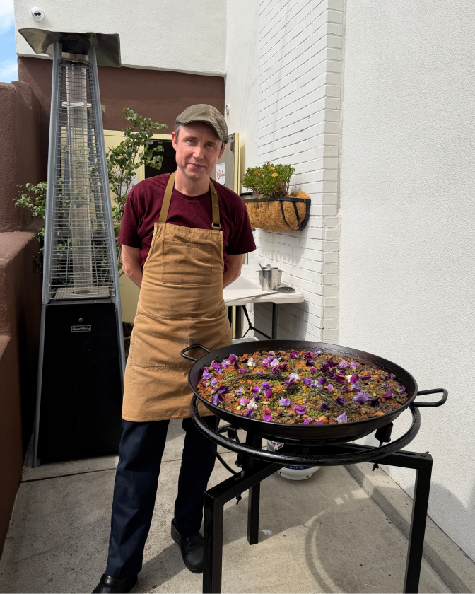 A man with a light skin tone, wearing a flat cap, maroon T-shirt and tan apron, stands behind a large black paella pan resting on a metal burner stand outdoors. The pan holds finished paella garnished with purple flowers and rosemary. A tall patio heater and a wall-mounted planter with greenery are visible behind him.