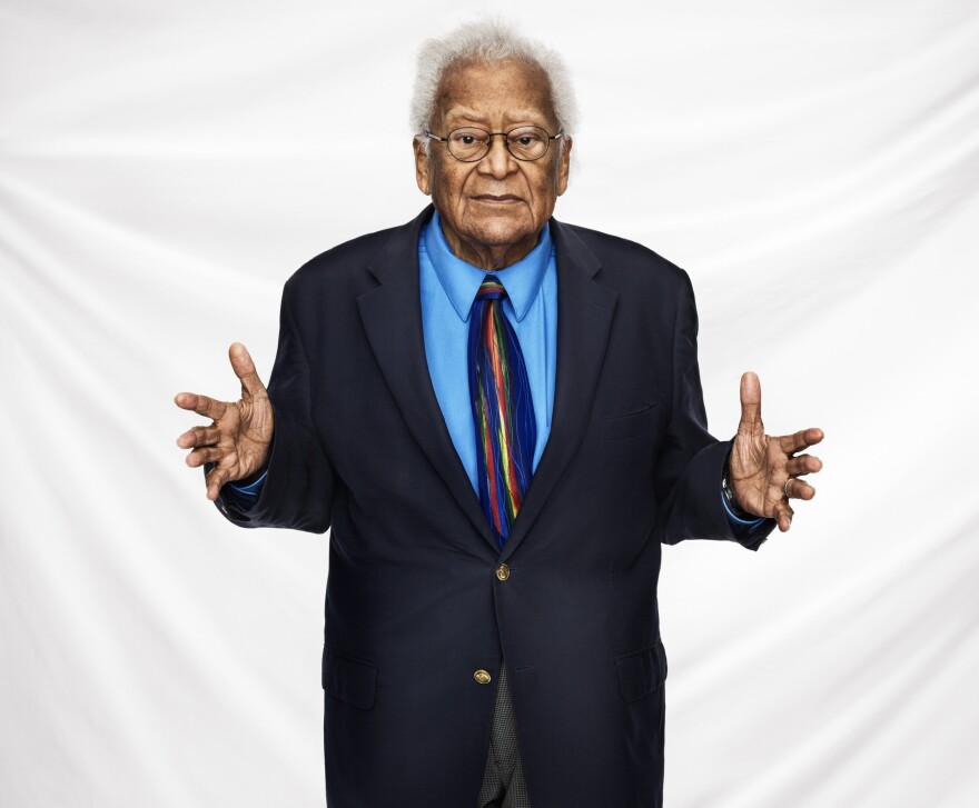 An older Black man stands against a white backdrop with his arms outstretched, palms open.