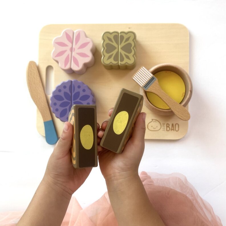 A pair of child hands holds a wooden toy shaped like a mooncake. In the background there are pink, purple and brown mooncakes.