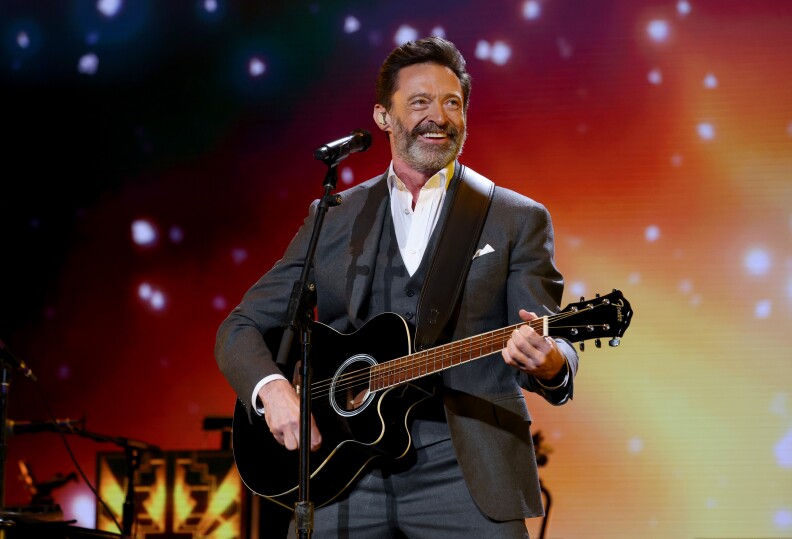 Hugh Jackman, a man in a suit playing an acoustic guitar on stage, stands in front a microphone stand. The background is lit with colorful lights and bokeh effects, creating a vibrant concert atmosphere.