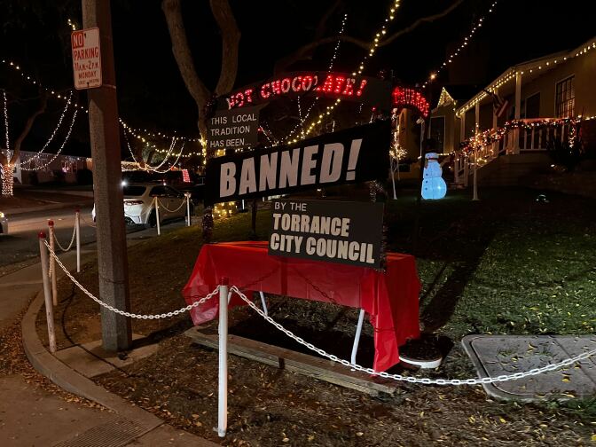 A small white foldable table, covered in a red tablecloth, is set up on a street corner outside of a home covered in holiday lights. A large black and white sign is affixed to the front of the table which reads "This local tradition has been BANNED! by the Torrance City Council"