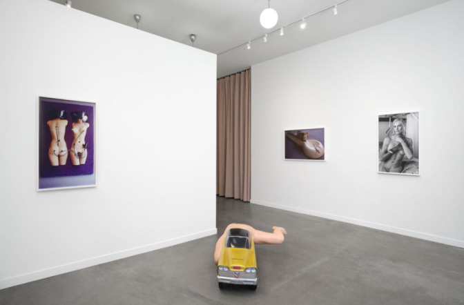 An art gallery with three provocative photographs on the wall, including a nude portrait. In the middle of the room is a sculpture of a half-car, half-human. 