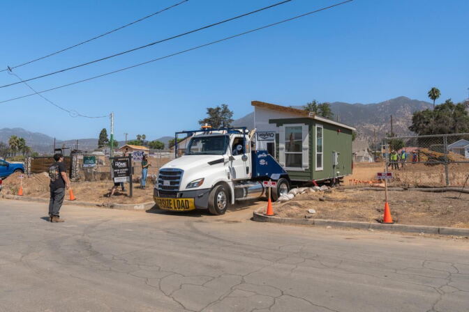 A truck with a small home on its trailer is parked in an empty lot, outlined against a clear blue sky.