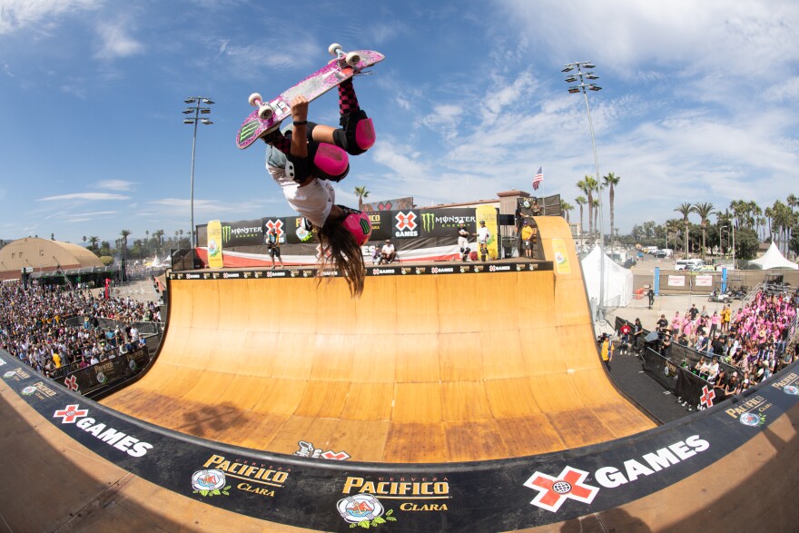 Skateboarder Arisa Trew performs during the Skateboard Vert Final at 2023 X Games.. She's flipped upside down, catching air above a half-pipe, wearing a pink helmet and pink protective gear.