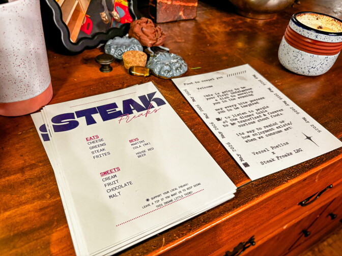A stack of Steak Freaks menus and a Vessel Poetics welcome card rest on a wooden dresser alongside clay pottery pieces, a candle, and other decorative objects.