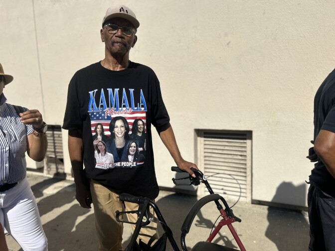 An older Black man wears a light grey baseball cap and black t-shirt that reads "Kamala Harris" in red and blue and features images of Harris in front of an American flag. He has a walker and stands in the sun in front of an off white wall. 