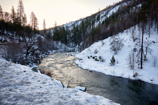 A stream cuts through a valley with snow-covered hills and pine trees on both sides.