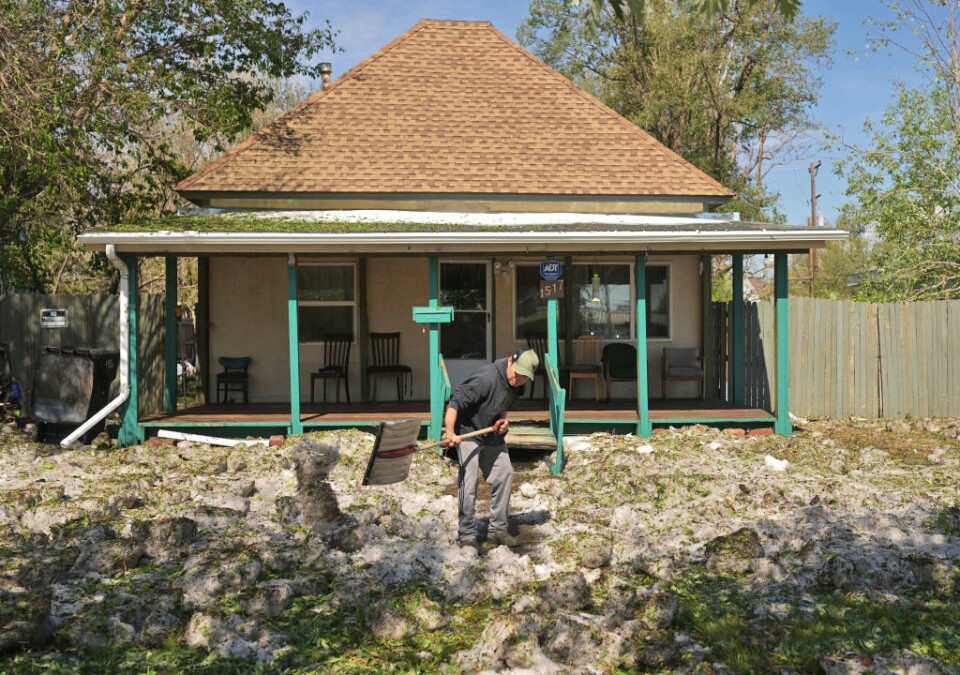 Photo of a man wearing a black top, grey jeans and a baseball cap holding a large shovel in front of a home with green columns and large porch