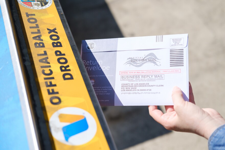 A ballot is being dropped in the slot at an official County of Los Angeles ballot drop box. Only the voter's hands are visible.
