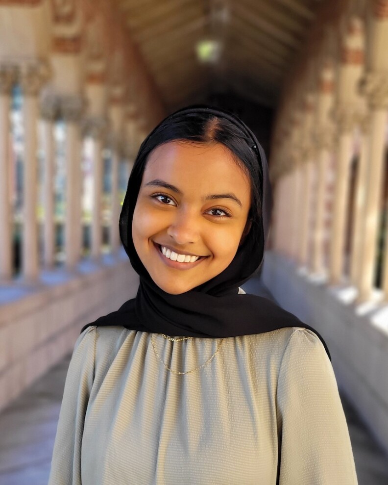 A female with medium skin tone smile towards the camera wearing a black headscarf and olive green top.