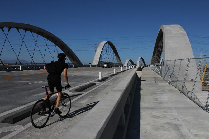 A person pedals a bicycle in a bike lane on a bridge. Large concrete arches border the roadway.