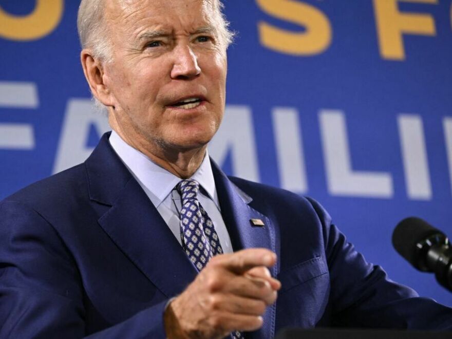 A man with light-tone skin and gray hair wears a dark blue suit and points his right hand in front of a microphone.