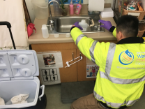 A person in a neon jacket and gloves kneels down and holds a bottle in a sink.