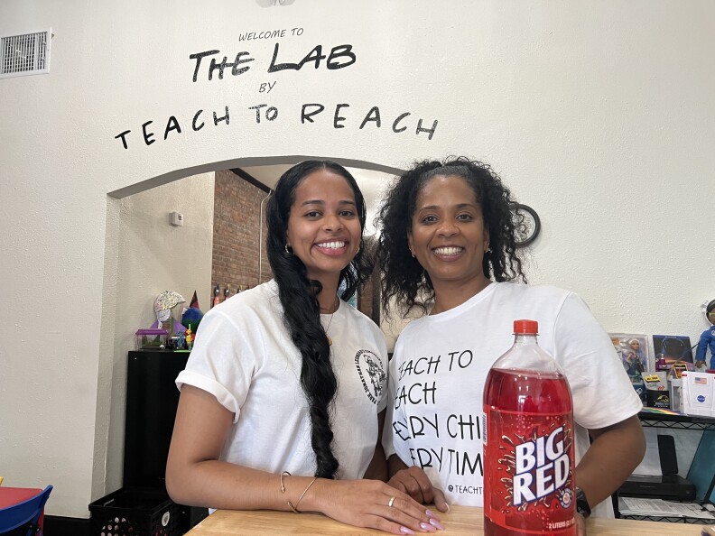 Two brown-skinned Black women in white shirts pose with a bottle of Big Red soda inside of a building. On the wall behind them is painted a message that reads "Welcome to The Lab by Teach to Reach."