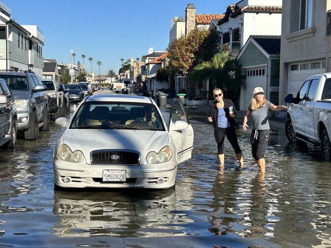 A car is stopped on a flooded residential street, where two women are wading through shin-deep water.