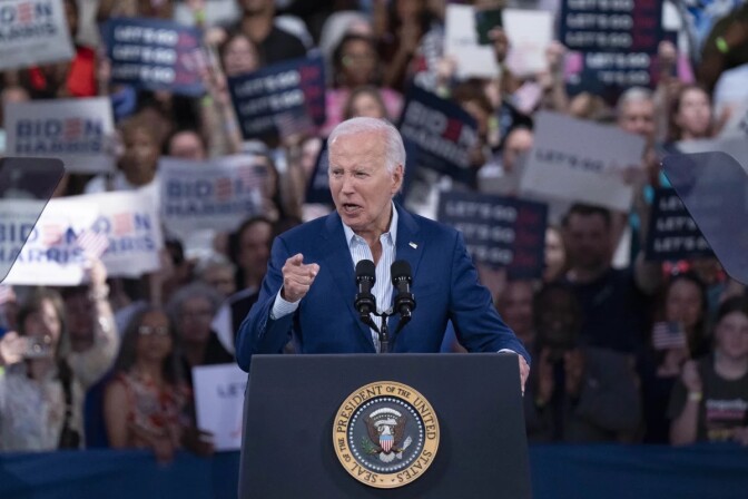A man with light-tone skin gestures at a lectern. A crowd is behind him. The lectern has the presidential seal.