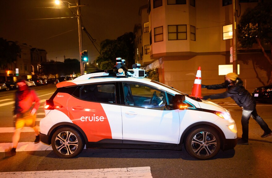 A person is placing an orange traffic cone on the hood of small car that has the word "cruise" on its side.