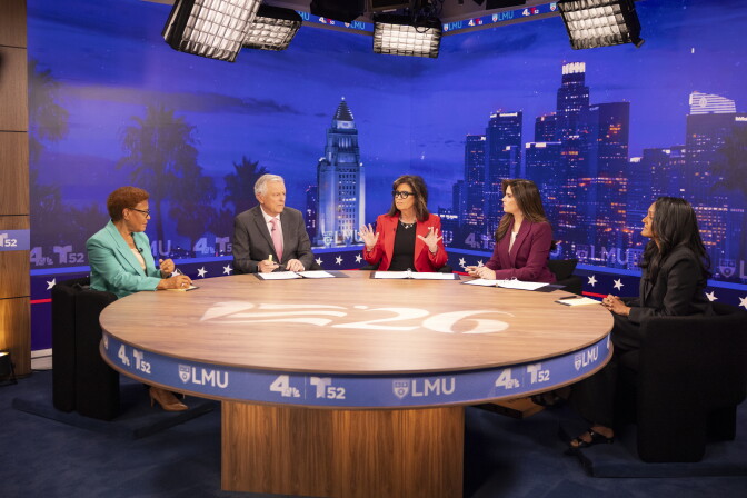 Five people sit at a round table in a TV studio with a Los Angeles skyline backdrop for a debate.