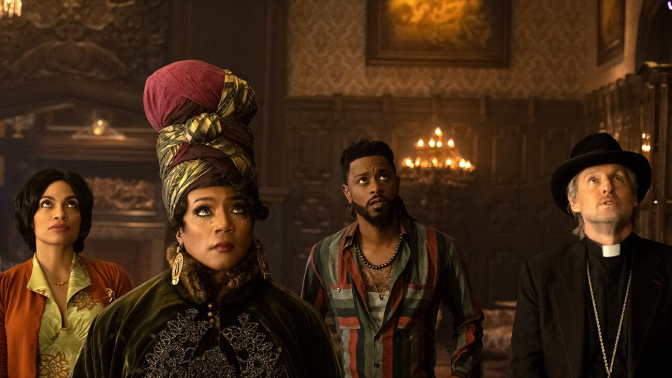Rosario Dawson, Tiffany Haddish, LaKeith Stanfield, and Owen Wilson stand in an ornate living room with carved wooden wall panels with candelabras hanging off those walls, washing the room in a dim golden glow with a slight haze of dust. They all stare up at something on the ceiling or high on the wall in front of them. Stanfield and Wilson look on seemingly worried while Dawson and Haddish look more unphased by what they are witnessing. Dawson wears a an orange sweater over a green collared shirt. Haddish wears a tall headscarf and heavy dark green cloak. Stanfield wears a green, red, and blue striped button down. Wilson wears a black fedora-like hat and priest suit. 