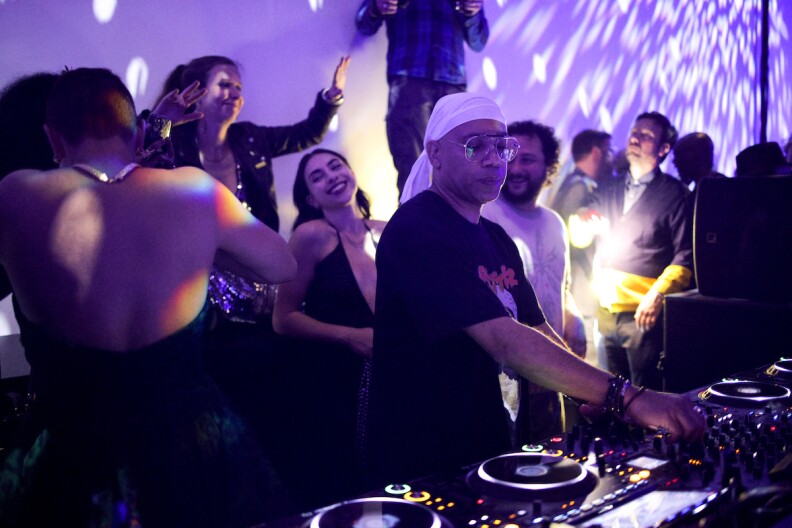 A man in glasses and a white head scarf DJs at a turntable spins tunes while several people bathed in purple light.