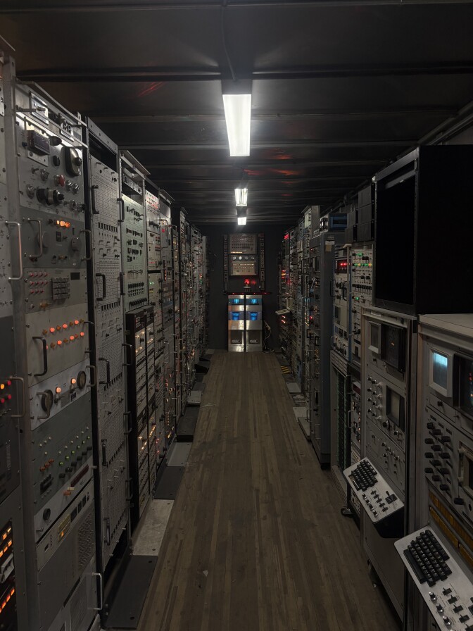 A narrow dark hallway with grey carpeting and wall-to-wall electronics with lights, buttons and knobs on either side. Strips of fluorescent lights line the dark ceiling.