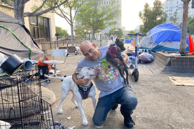 A smiling man wearing a gray t shirt and blue jeans is holding two dogs while kneeling on the sidewalk in the middle of an encampment for unhoused people. 