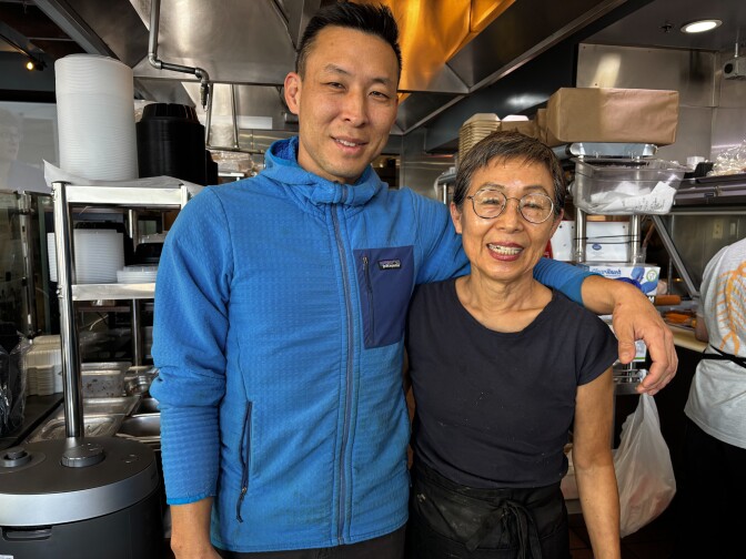 Two people with medium dark skin standing side by side inside the restaurant kitchen, smiling at the camera. The man on the left wears a blue jacket; the woman on the right wears glasses and a black shirt, with stainless steel kitchen equipment behind them.