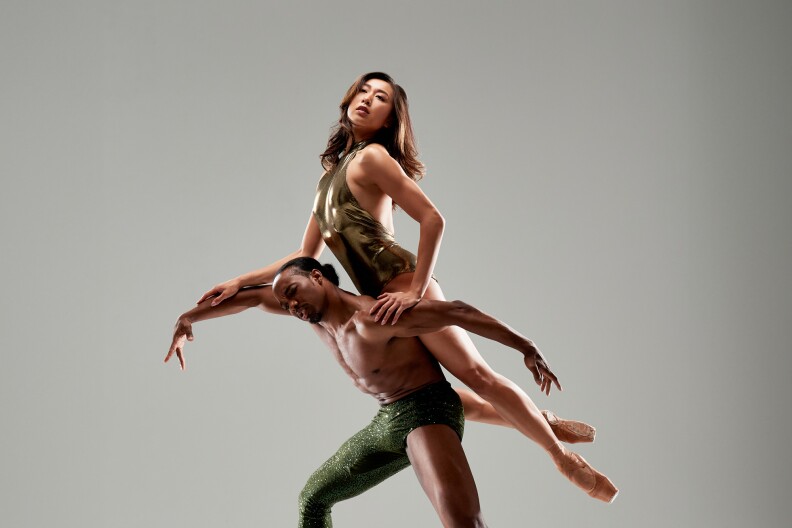 Two ballet dancers pose in a studio with a gray backdrop. The man wearing sparkly green pants is poses with his legs and arms outstretched, supporting the woman in his back as she extends her legs while wearing pointe shoes.